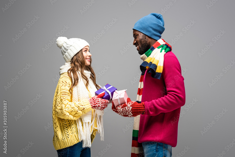 side view of happy interracial couple in winter outfit holding presents on grey background side view of happy interracial couple in winter outfit holding presents on grey background