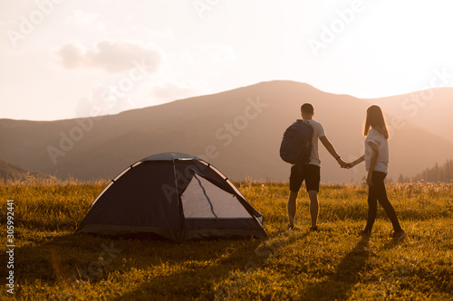 Back view of young couple walking with backpacks at autumn mountains sunset near tent. Camping, hiking.
