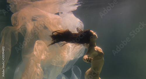 Beautiful girl swims underwater with long hair. Blue or gold background like gold. The atmosphere of a fairy tale or magic. Diving under the water with a shiny cloth