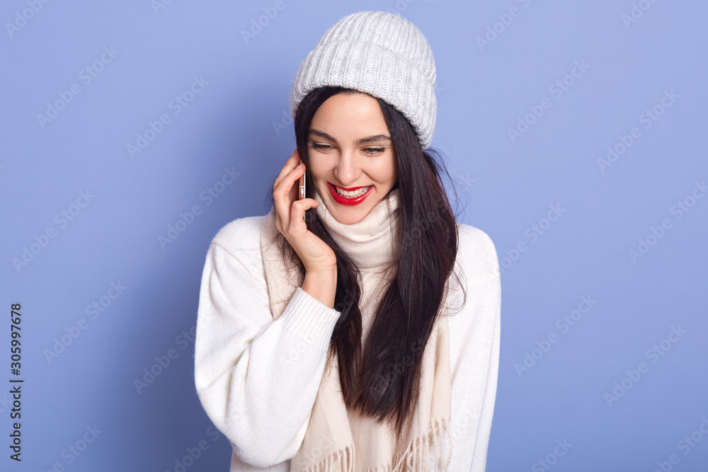 Happy young woman talking to her friend via smart phone isolated over blue background, lady with red lips dresses white warm clothes, brunette girl having joyful facial expression. People concept. Happy young woman talking to her friend via smart phone isolated over blue background, lady with red lips dresses white warm clothes, brunette girl having joyful facial expression. People concept.