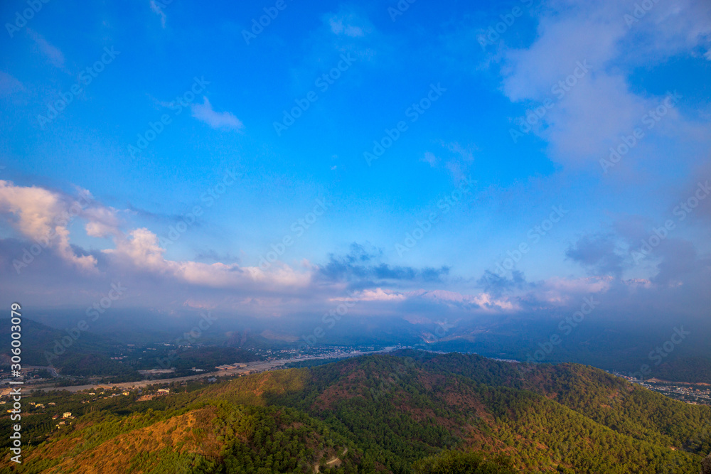Amazing view from the top of cliff  to the city located on foot of mountains. The rock under fairytale blue sky with the clouds.  Beautiful mountain landscape. Panorama.