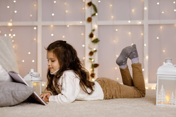 Happy little child girl is reading book on the background with lights. Merry Christmas