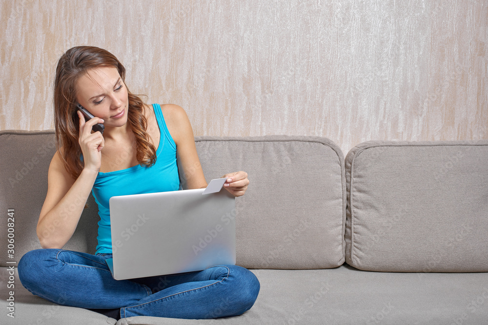 Beautiful woman wearing cyan top with thin shoulder straps and jeans sitting cross-legged on cushions sofa looking at plastic card in her hand and talking on the phone, laptop on lap. Beautiful woman wearing cyan top with thin shoulder straps and jeans sitting cross-legged on cushions sofa looking at plastic card in her hand and talking on the phone, laptop on lap.