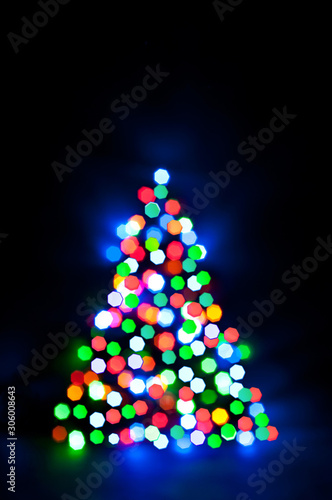 Christmas fairy lights are defocused in the shape of a fir tree, giving a blurry effect. Dark background for design.