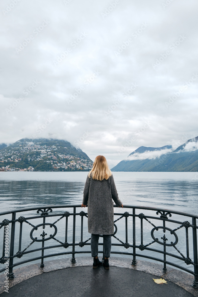 Beautiful caucasian woman stands on pier in Lugano. Girl in travel. Lake Lugano, southern slope of Alps. Landscape in Switzerland. Amazing scenic outdoors view. Canton of Ticino. Adventure lifestyle