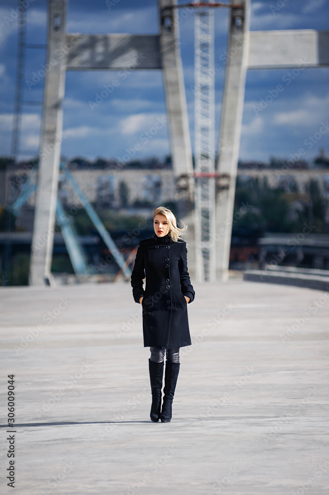 A blonde in a black classic coat walks on unfinished bridges over the river in the fall. Autumn photo shoot in the city A blonde in a black classic coat walks on unfinished bridges over the river in the fall. Autumn photo shoot in the city