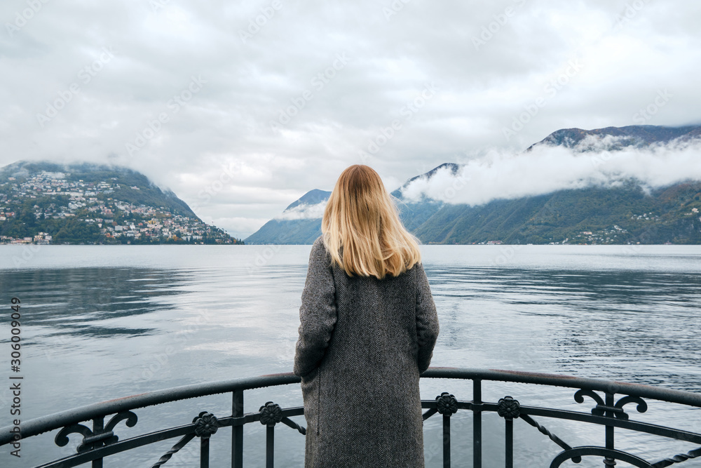 Beautiful caucasian woman stands on pier in Lugano. Girl in travel. Lake Lugano, southern slope of Alps. Landscape in Switzerland. Amazing scenic outdoors view. Canton of Ticino. Adventure lifestyle Beautiful caucasian woman stands on pier in Lugano. Girl in travel. Lake Lugano, southern slope of Alps. Landscape in Switzerland. Amazing scenic outdoors view. Canton of Ticino. Adventure lifestyle