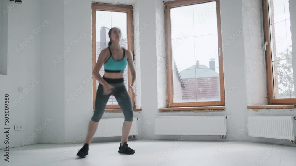 Young fit brunette girl with pigtail in blue top, gray leggings, sneakers does exercises in bright room. Doing sports and fitness training at home. Healthy lifestyle. Squats, jumps, female jumping Young fit brunette girl with pigtail in blue top, gray leggings, sneakers does exercises in bright room. Doing sports and fitness training at home. Healthy lifestyle. Squats, jumps, female jumping