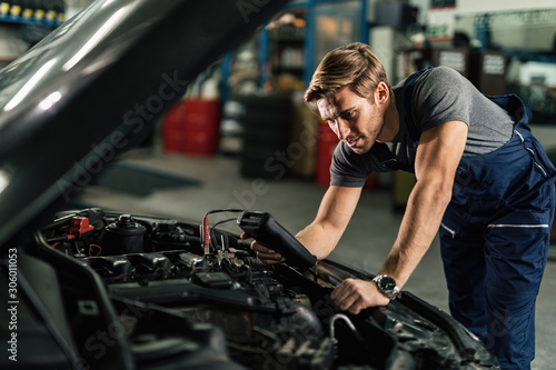 Young mechanic with diagnostic tool analyzing car engine problem in auto repair shop.