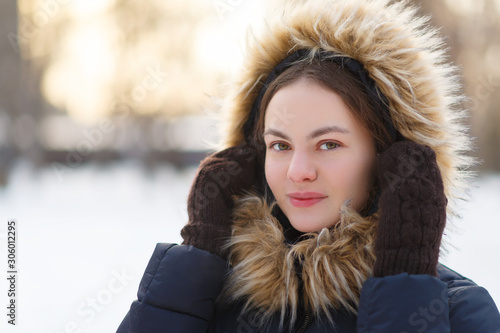 winter portrait of young attractive smiling girl in downy winter jacket with hood and knitted mittens