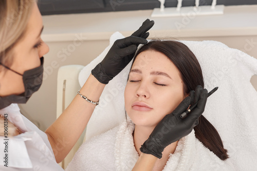 Close-up Of Surgeon Hands Drawing Correction Lines On Woman Face With Marker. Doctor's hand in black surgical glove marking women's face for cosmetic injections.