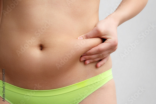 the woman's hand holds a fold of excess fat on her stomach. on a gray background, front view