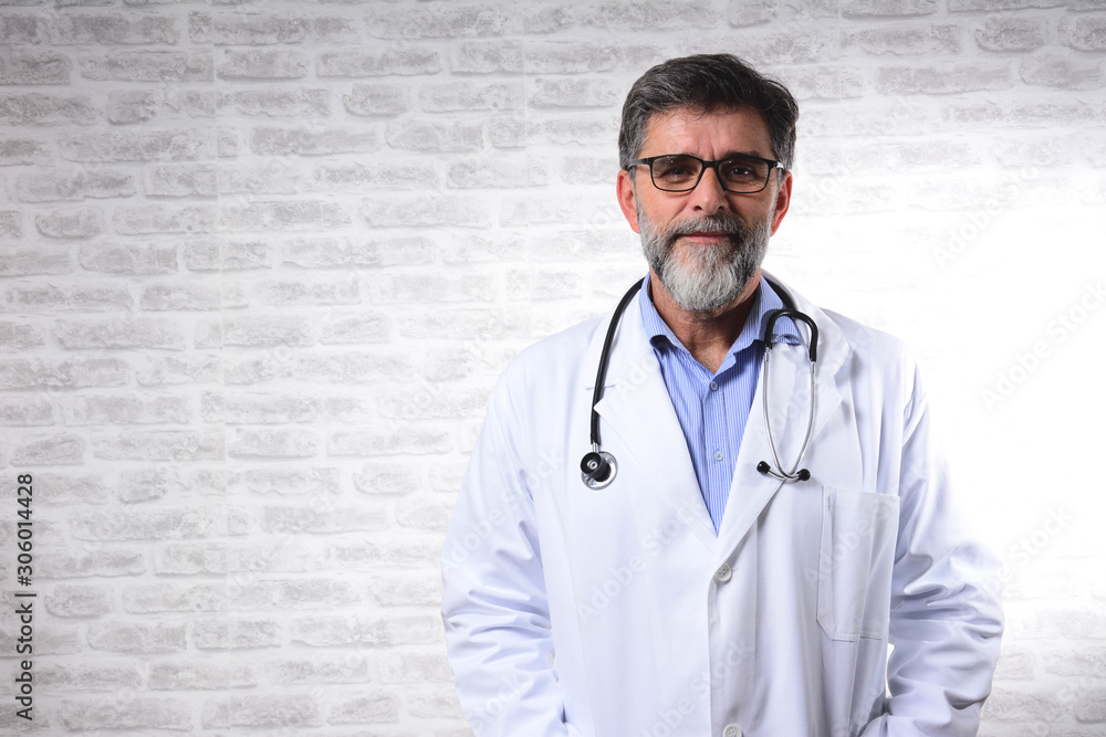 Portrait of an attractive senior male doctor in white coat. Portrait of glad smiling doctor in white uniform standing on gray brick wall background. Stethoscope or phonendoscope Portrait of an attractive senior male doctor in white coat. Portrait of glad smiling doctor in white uniform standing on gray brick wall background. Stethoscope or phonendoscope