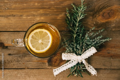 Crystal mug filled with green tea with a slice of lemon inside of the beverage next to aromatic cooking herbs as thyme and rosemary tied up with a lace bow in a wooden background