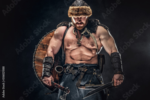 Tablou pe pânză Severe barbarian in warrior clothes, posing on a dark background.