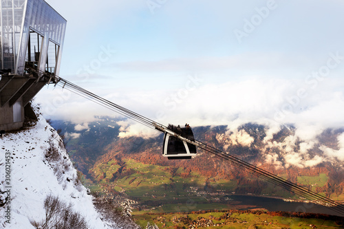 Photo of a cabrio funicular, departing from the Stanserhorn mountain station in Switzerland. Presence of snow on the top of the mountains, and green meadows in the valley, illuminated by the light of 