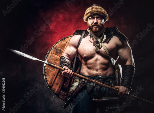 Serious Viking clad in light armor with a shield behind his back holds a spear. Posing on a dark background with red light