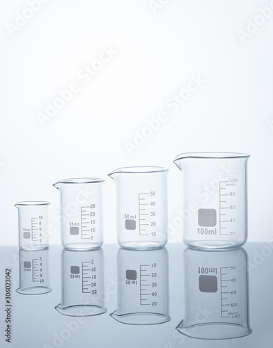 Four empty measuring beakers sitting on a mirror blue surface, glass lab containers standing in gradual order on a table, transparent light through laboratory flasks, precise comparing measurements 