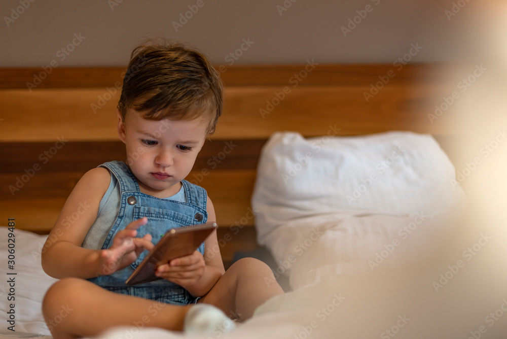 Fototapeta premium Wow, I like that phone. Baby with smartphone. Little boy sitting in bed and playing with mobile phone. Calling my mom. Cute little baby holds mobile phone in his hands 