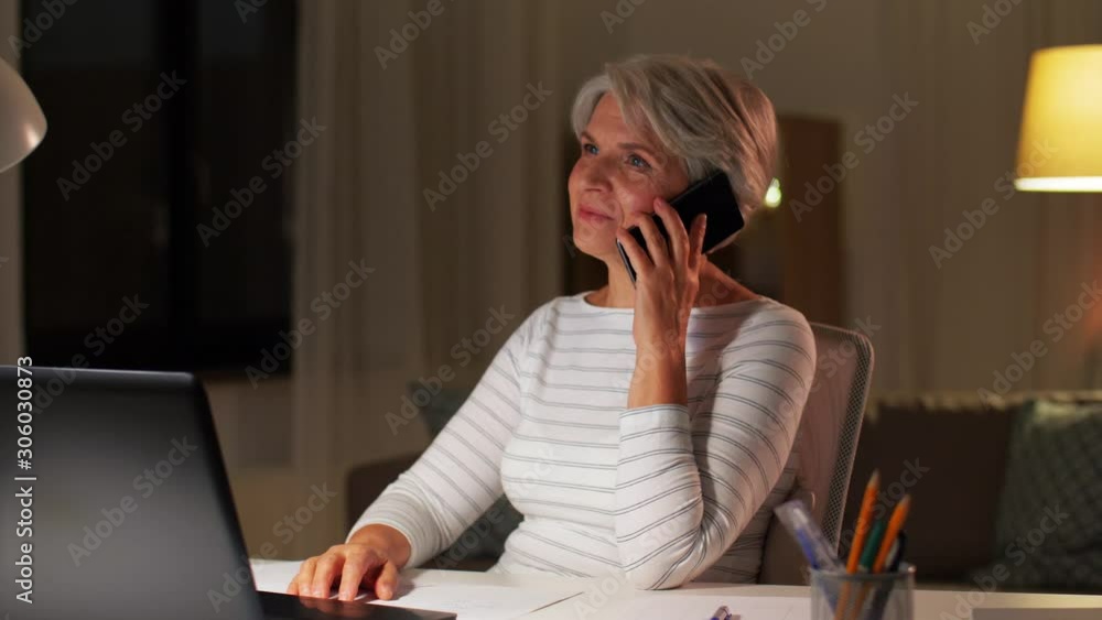 technology, old age and communication concept - senior woman with laptop calling on smartphone at home in evening technology, old age and communication concept - senior woman with laptop calling on smartphone at home in evening