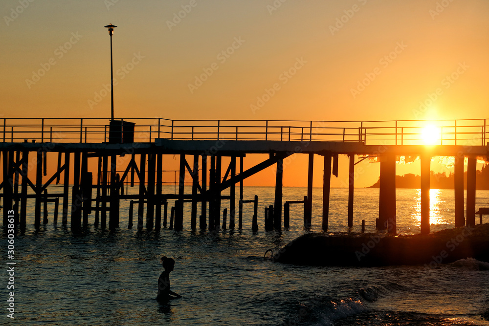 Silhouette of a girl who is swimming in the sea near the pier during sunset.