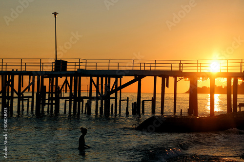 Silhouette of a girl who is swimming in the sea near the pier during sunset. © Nadzeya
