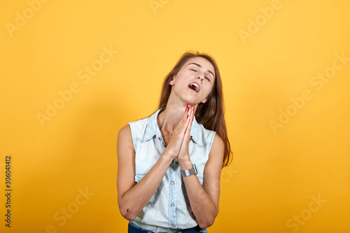 Attractive caucasian young woman in blue denim shirt praying, keeping hands together isolated on orange background in studio. People sincere emotions, lifestyle concept.