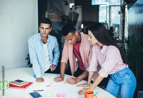 Portrait of serious Spanish male entrepreneur looking at camera while colleagues writing ideas during briefing time, clever diverse team cooperating on startup project near office desktop