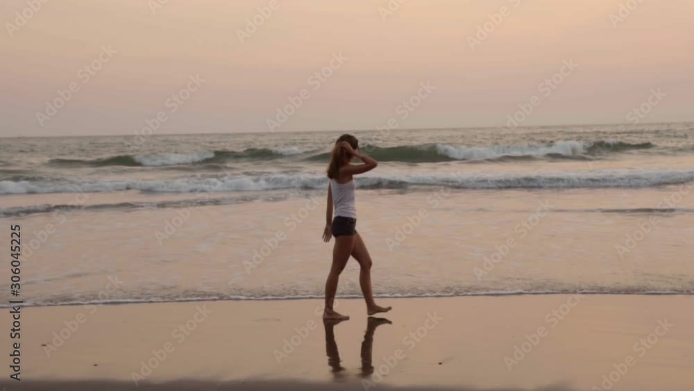 Side view slow motion shot of young slim woman walking barefoot along ocean shore at sunset and then raising leg forward and making water splash Side view slow motion shot of young slim woman walking barefoot along ocean shore at sunset and then raising leg forward and making water splash