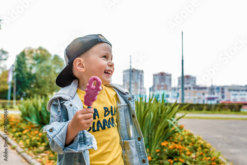 Happy little boy 3-5 years old  in summer in city park  smiles  rejoices  eats ice cream on stick. Autumn clothes. Emotions of pleasure and relaxation. Laughs has fun. Free space for copy text.