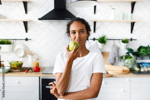 Charming african girl is eating an green apple in the kitchen