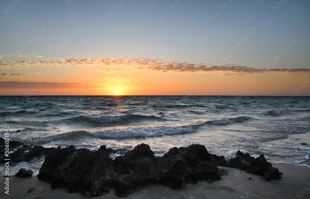 Sun sets on horizon over bay with blue sky, gentle waves and rocks in foreground.