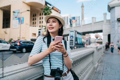 smiling beautiful woman tourist using smartphone in pedestrian zone of city during summer time. happy girl traveler searching direction online map on cellphone. female photographer relax on sidewalk