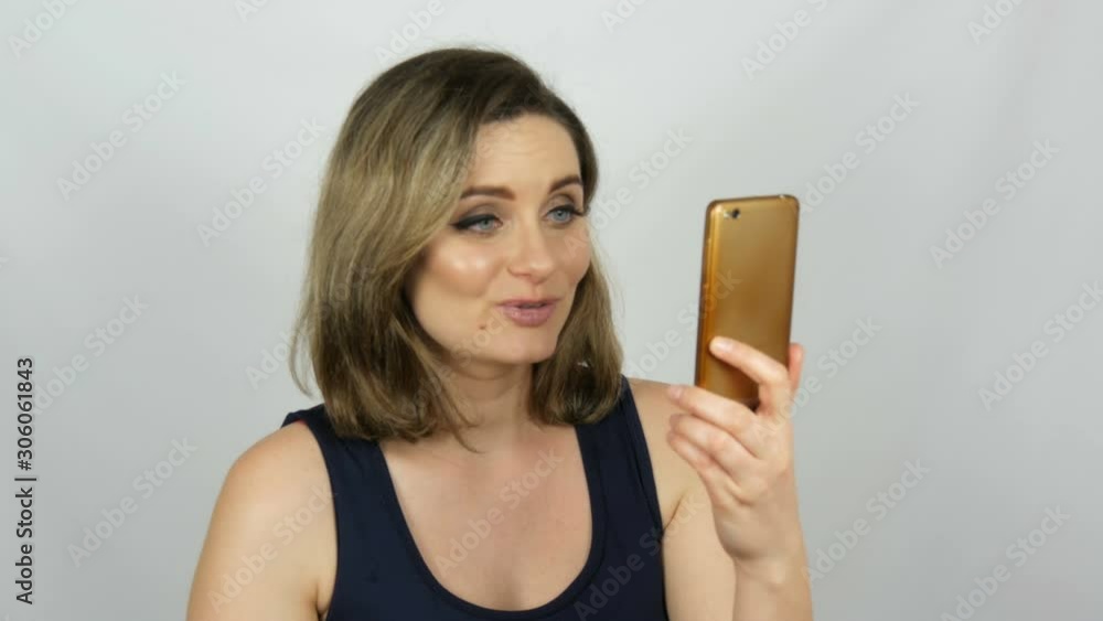 Portrait of a beautiful young woman who is posing making a selfie and communicating and looking into a smartphone that is holding in her hands on a white background in the studio. Portrait of a beautiful young woman who is posing making a selfie and communicating and looking into a smartphone that is holding in her hands on a white background in the studio.