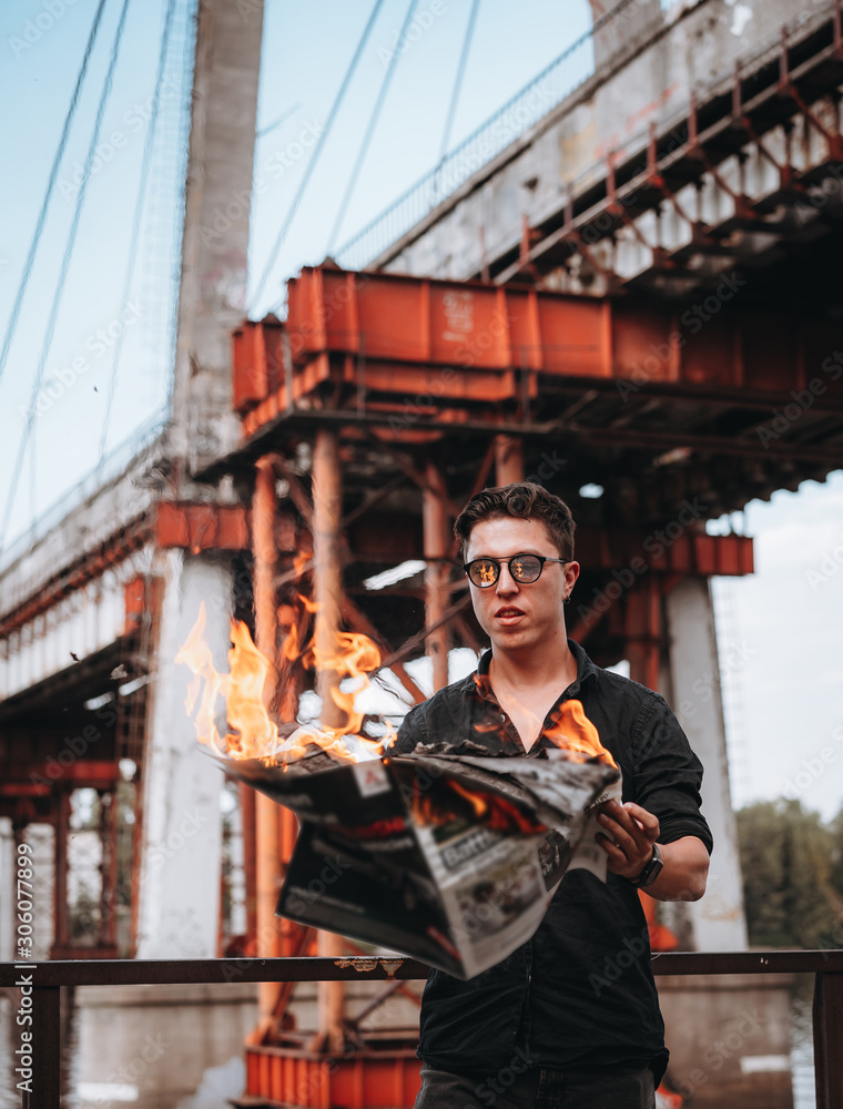 Guy reads a burning newspaper, in the background a bridge