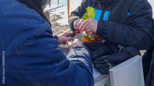 Two men in warm winter clothes use a digital multimeter to check the interface cable connector while outdoors in winter.