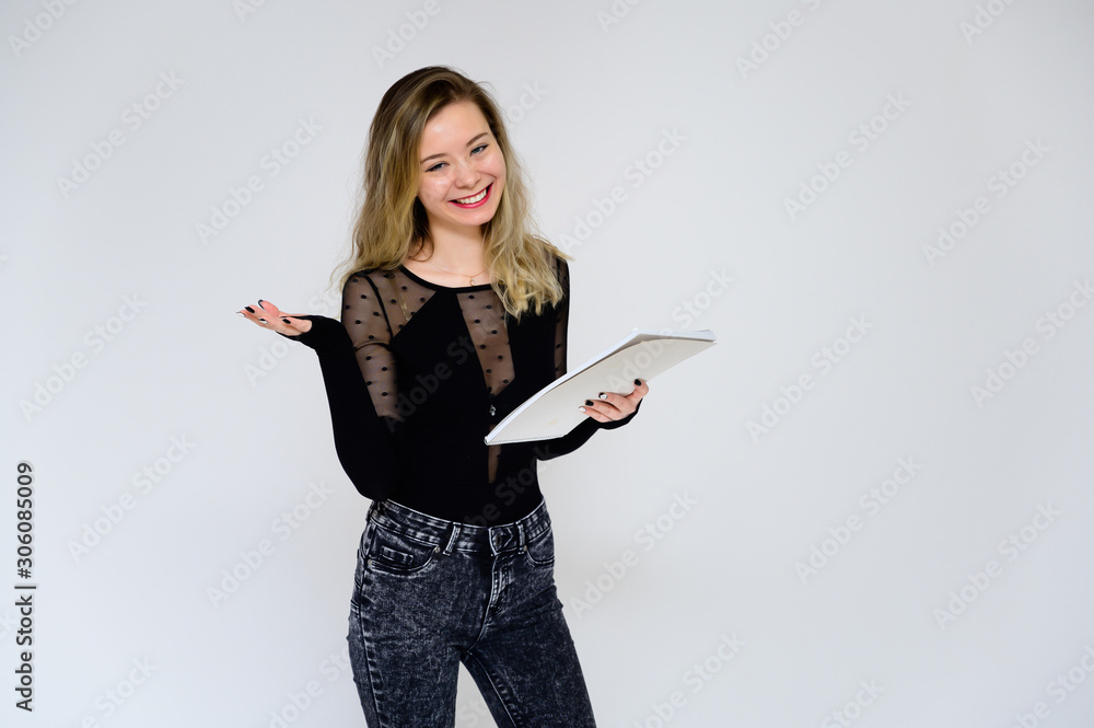 Concept of a woman talking to the camera. A photo of a pretty smiling girl with curly hair in a black T-shirt on a white background is standing right in front of the camera with a folder in her hands. Concept of a woman talking to the camera. A photo of a pretty smiling girl with curly hair in a black T-shirt on a white background is standing right in front of the camera with a folder in her hands.