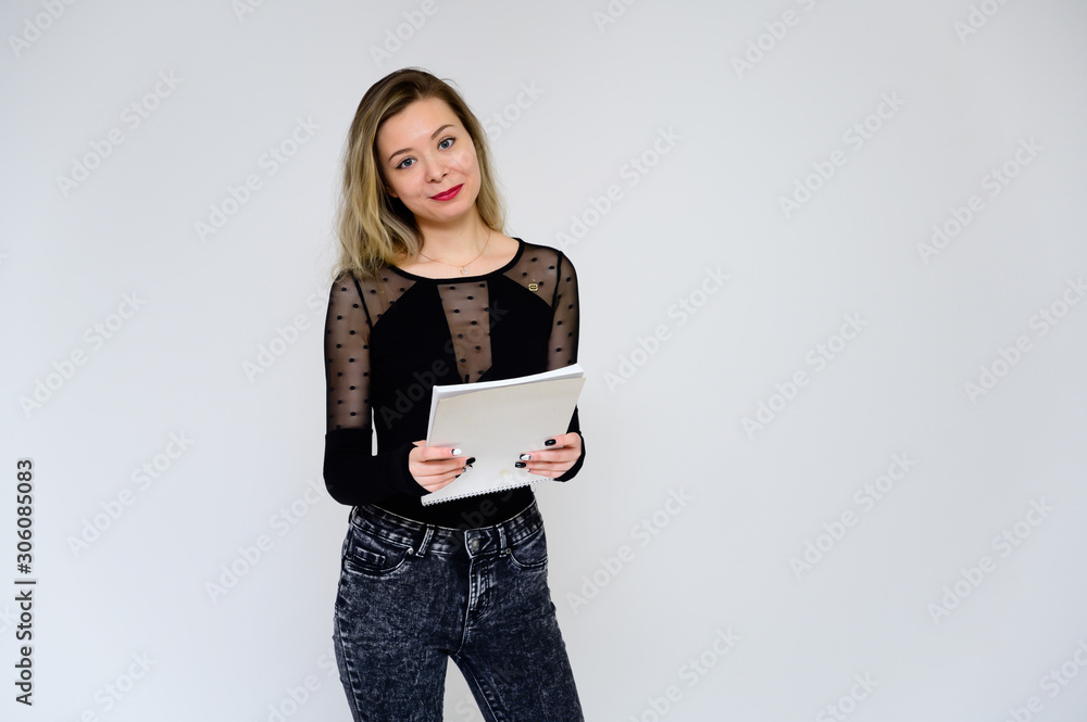 Concept of a woman talking to the camera. A photo of a pretty smiling girl with curly hair in a black T-shirt on a white background is standing right in front of the camera with a folder in her hands. Concept of a woman talking to the camera. A photo of a pretty smiling girl with curly hair in a black T-shirt on a white background is standing right in front of the camera with a folder in her hands.