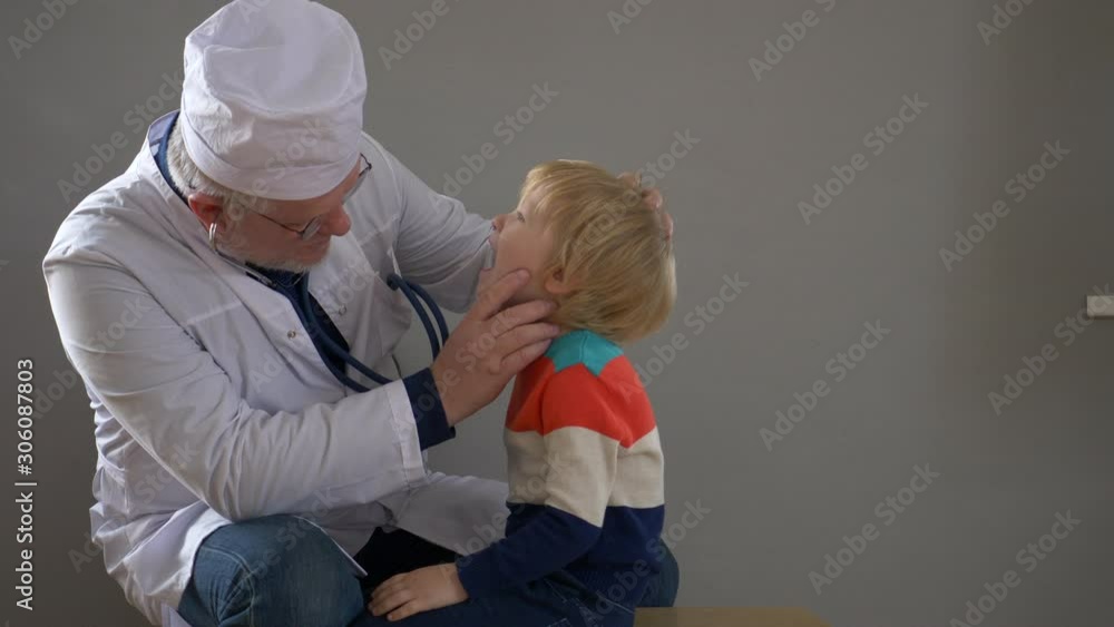 elderly doctor with gray hair and glasses in white robe and cap examines cute little blonde boy with stethoscope. doctor examines throat and listens to lungs. Child trusts medical man elderly doctor with gray hair and glasses in white robe and cap examines cute little blonde boy with stethoscope. doctor examines throat and listens to lungs. Child trusts medical man