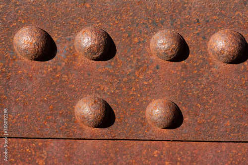 Six sunny rusty bolts head of a steel footbridge and their shadow on a rusty beam. Centered at the top of the image.