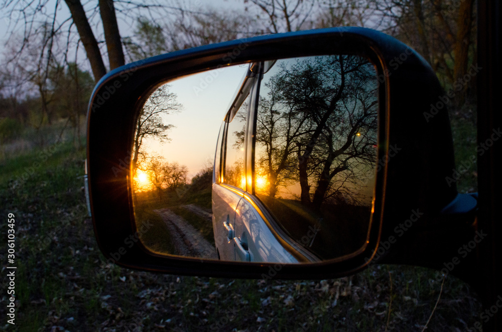 Side rearview mirror of the car with the reflection of the beautiful sunset in the forest. Close up. View of the sun through the autumn trees. Concept of family road trips and travel.