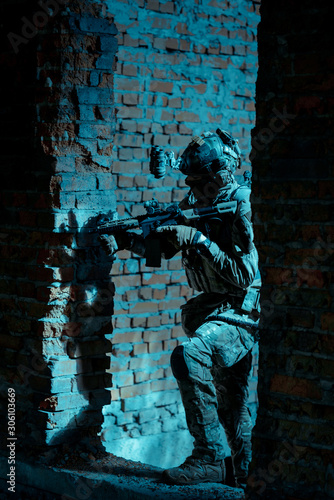 Man in uniform with machine gun and night-vision device move between two broken walls of building. Airsoft soldier in night building
