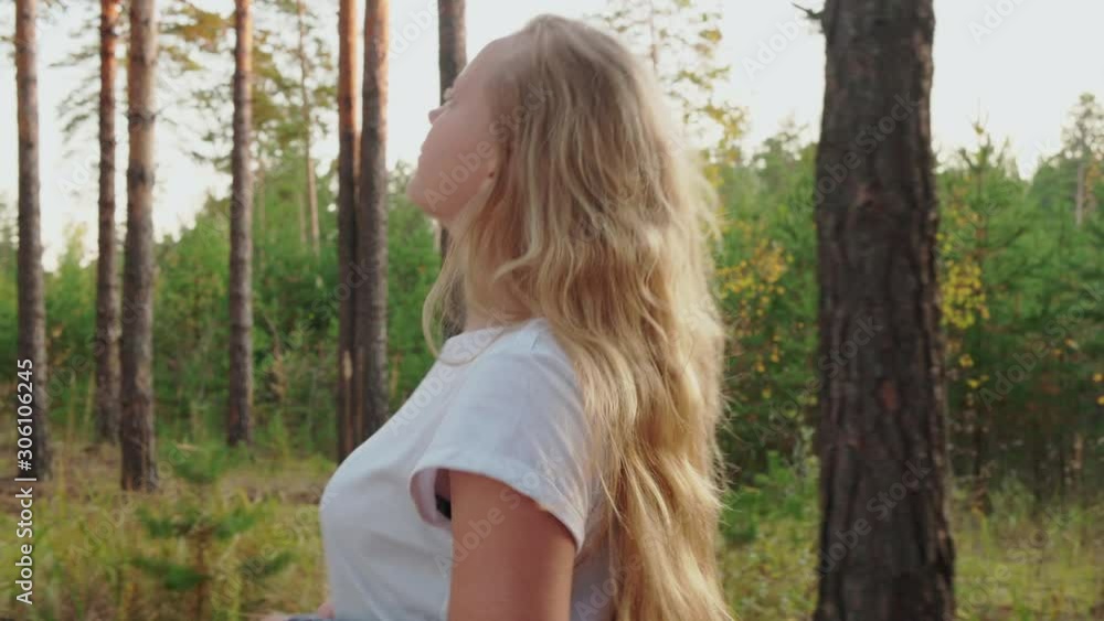 Side view of beautiful girl walking in pine forest. Adorable blonde girl in white t-shirt and checkered shirt walking in beautiful green forest and looking at pine trees during daytime Side view of beautiful girl walking in pine forest. Adorable blonde girl in white t-shirt and checkered shirt walking in beautiful green forest and looking at pine trees during daytime
