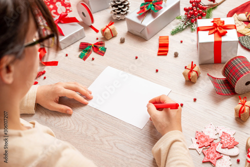 Girl writes Christmas and New Year greetings. Blank paper to add greeting card text. Mockup. A view over shoulder at a table full of gifts and decorations.