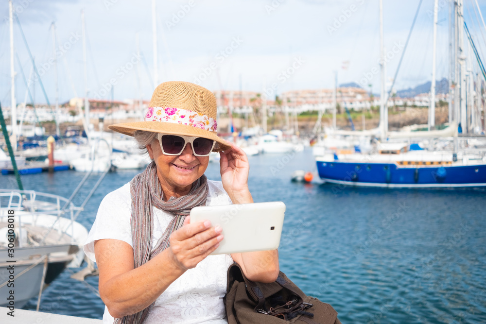 An elderly woman with gray hairs takes a selfie with the scenery with her tablet. A small port with yachts and sailboats. Cloudy and windy day