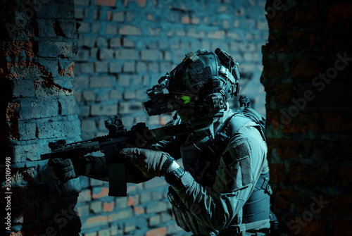Man in uniform with machine gun and turned on night vision device move between brick wall. Closeup airsoft soldier with green light on face in night