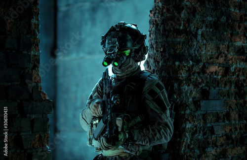 Man in uniform with machine gun and turned on night vision device stand between two brick walls. Airsoft soldier with green light on face in night