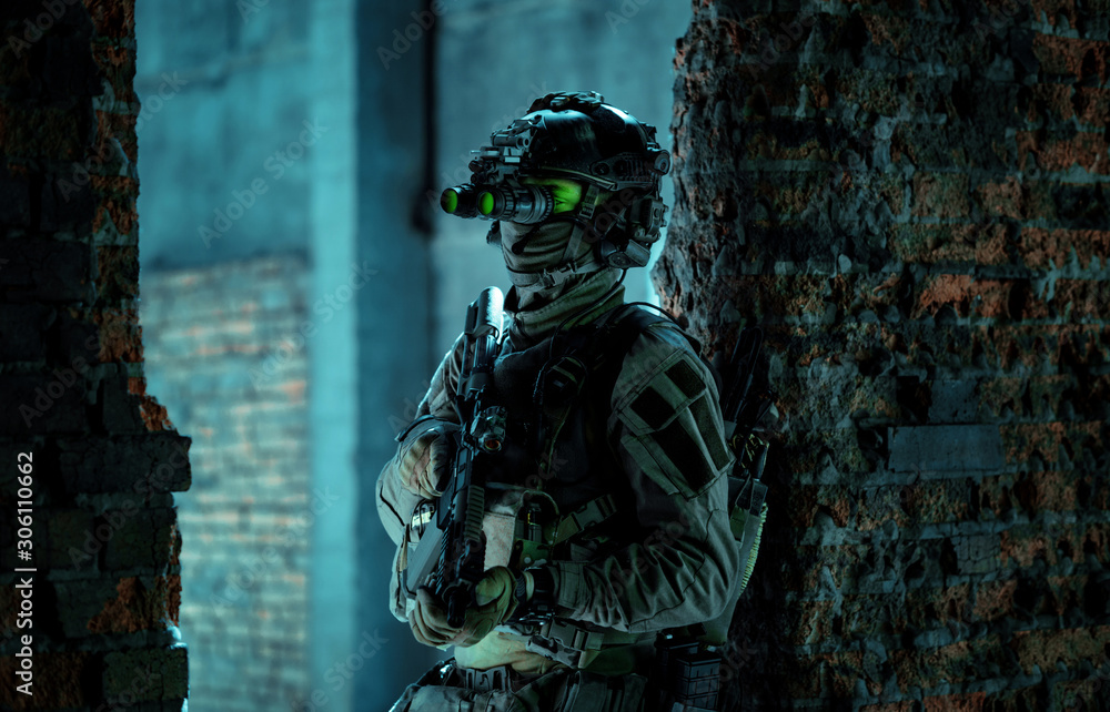 Man in uniform with machine gun and turned on night vision device stand between two brick walls. Airsoft soldier with green light on face in night Man in uniform with machine gun and turned on night vision device stand between two brick walls. Airsoft soldier with green light on face in night