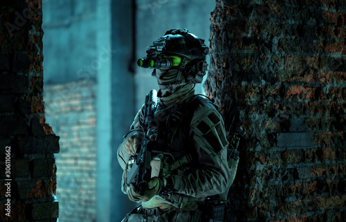 Man in uniform with machine gun and turned on night vision device stand between two brick walls. Airsoft soldier with green light on face in night