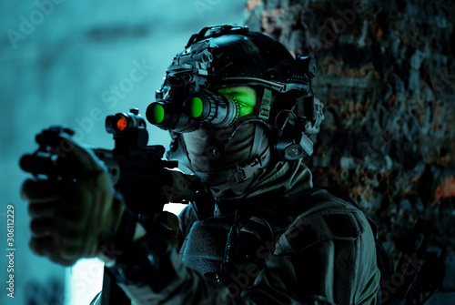 Man in uniform with machine gun and turned on night vision device beside brick wall. Closeup airsoft soldier with green light on face in night